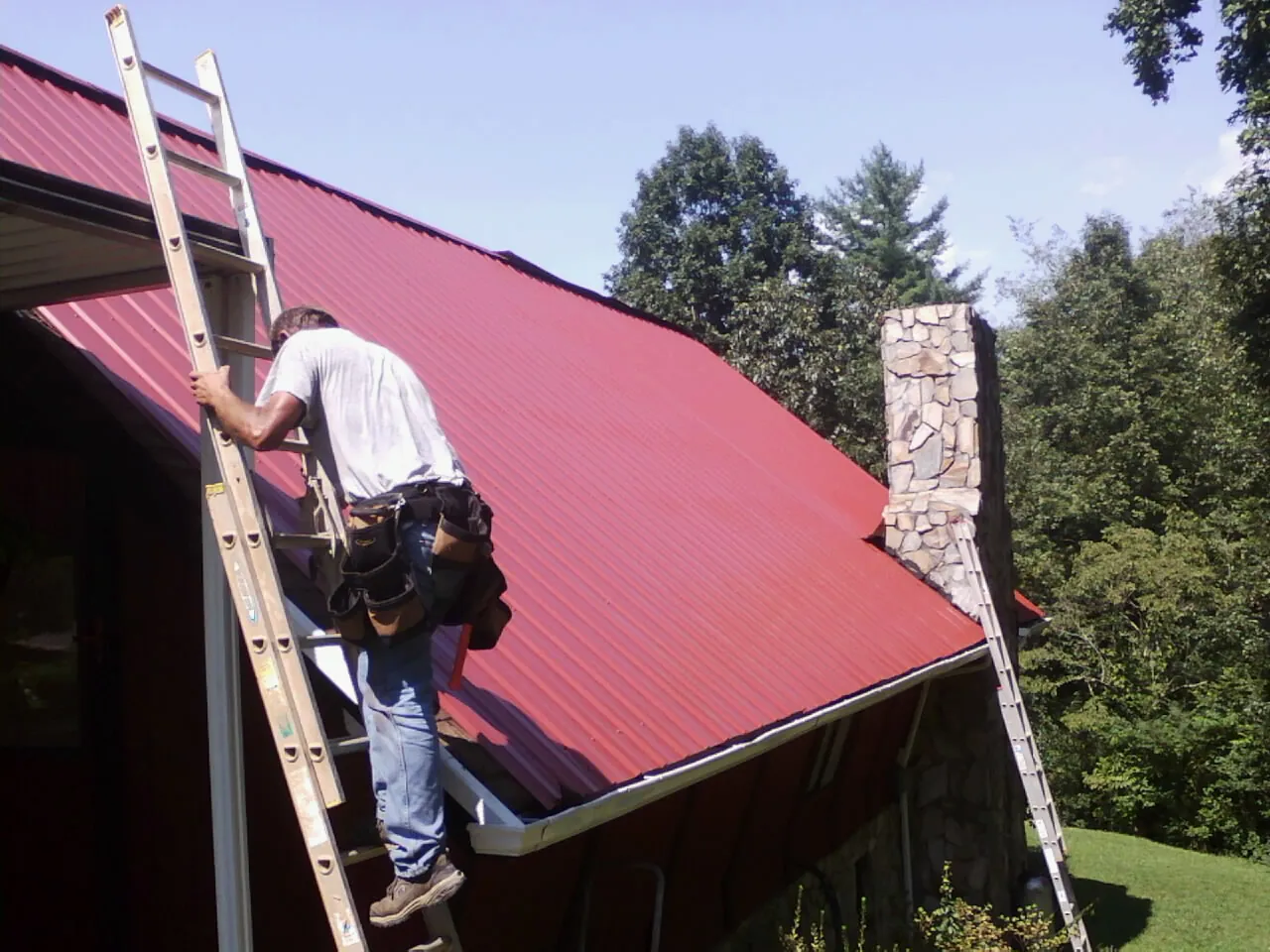 Roofer on a ladder beside a red metal roof and stone chimney — Soffit & Fascia Repair by Roofing Brothers in Stevenson Ranch